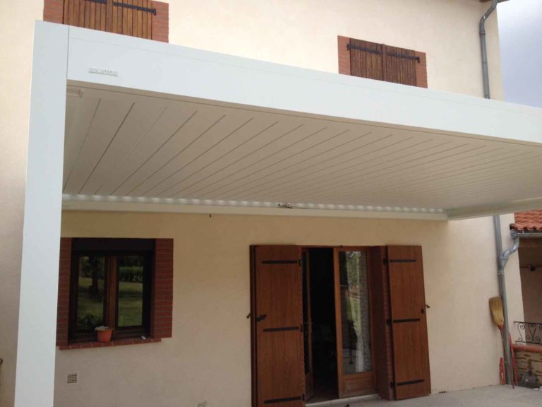 Pergola blanche à lames orientables contre une façade à volets bois