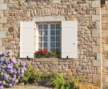 Fenêtre avec volets battants blancs ouverts sur une façade en pierre, encadrée de fleurs colorées.