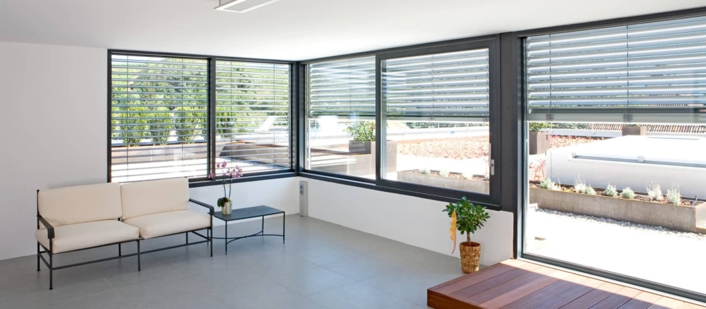 Intérieur moderne et lumineux avec de grandes baies vitrées et stores extérieurs, offrant une vue sur un jardin aménagé.