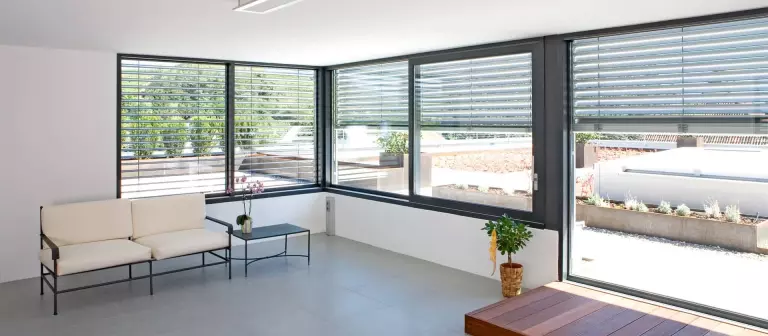 Intérieur moderne et lumineux avec de grandes baies vitrées et stores extérieurs, offrant une vue sur un jardin aménagé.
