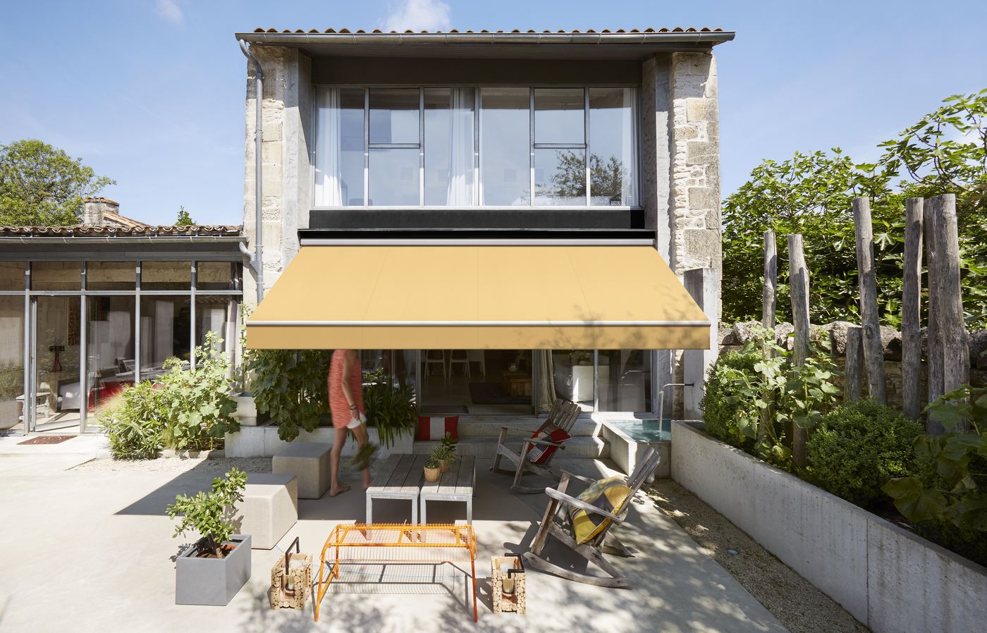Store banne à toile Dickson jaune sur une maison en pierre avec terrasse fleurie